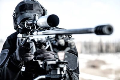 Sniper of police special operations tactical group in black uniforms, ballistic glasses and headphones, aiming with telescopic optical sight on sniper rifle mounted on tripod, over shoulder back view